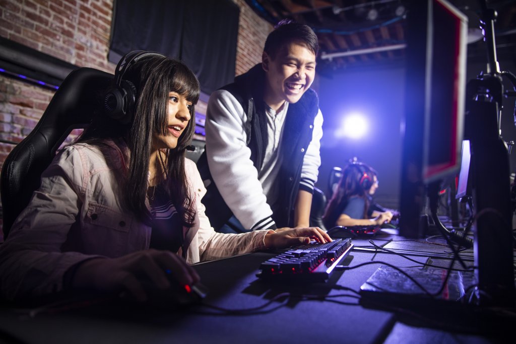 Jóvenes estudiantes universitarios divirtiéndose, riendo y jugando esports en estaciones de computadoras con monitores, teclados y ratones