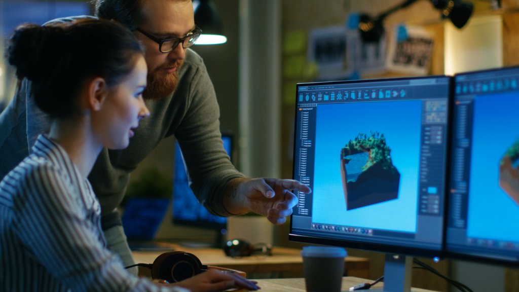 Female Game Developer Has Discussion with Male Project Manager While Working on a Game Level on Her Personal Computer with Two Displays. They Work in a Modern Loft Office Creative Environment.