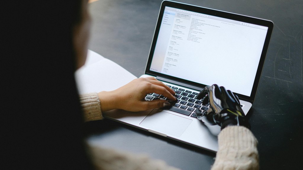 A woman possesses a human hand on her left side and a robotic hand on her right, working in tandem on a laptop.